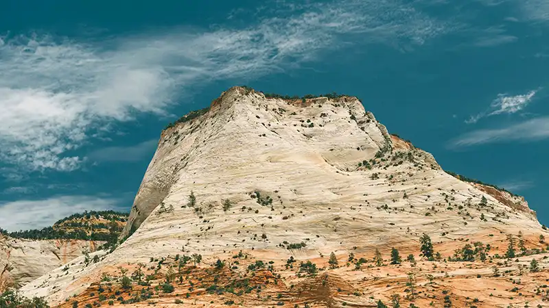 A large, light-colored sandstone mountain dominates the frame under a dramatic blue sky with wispy clouds. The mountain's face is a mix of off-white and light tan, marked by horizontal striations and dotted with small green trees and shrubs. The base of the mountain is a darker reddish-orange, suggesting a different rock layer or soil. To the left, another, darker mountain peak is partially visible. The overall impression is one of grandeur and rugged natural beauty.